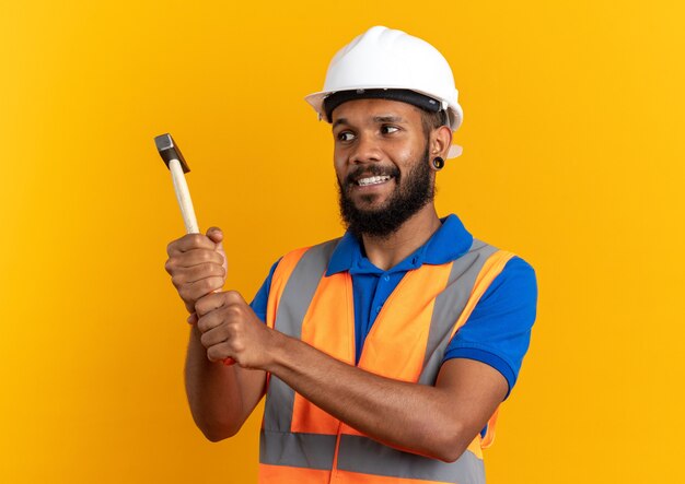 Sonriente joven constructor hombre en uniforme con casco de seguridad sosteniendo y mirando el martillo aislado en la pared naranja con espacio de copia