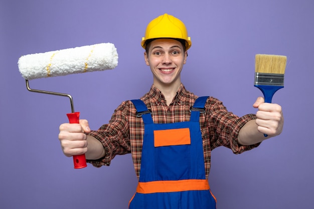 Sonriente joven constructor masculino vistiendo uniforme sosteniendo pincel con cepillo de rodillo