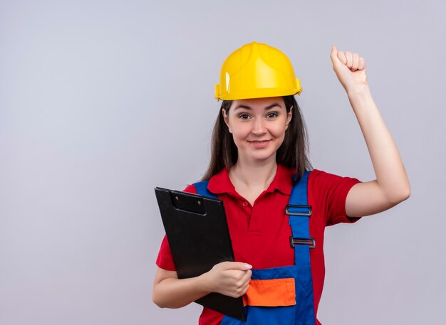 Sonriente joven constructor sostiene el portapapeles y levanta el puño sobre fondo blanco aislado con espacio de copia