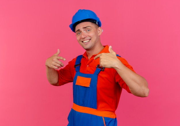 Sonriente joven constructor con uniforme y casco de seguridad apunta a sí mismo en rosa