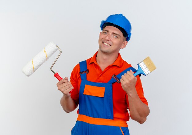 Sonriente joven constructor con uniforme y casco de seguridad con rodillo de pintura con pincel en blanco