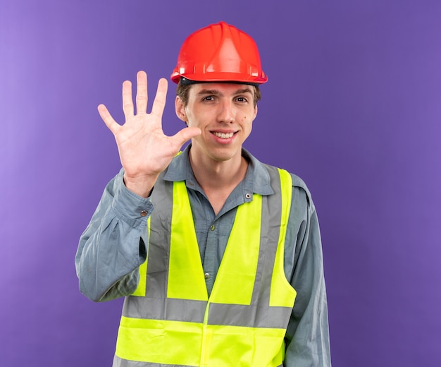 Sonriente joven constructor en uniforme mostrando cinco aislados en la pared azul