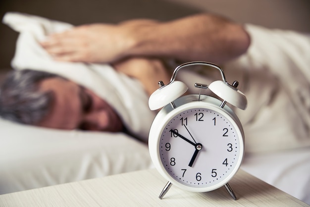 Sonriente joven cubriendo las orejas con almohada mientras mira el despertador en la cama. Hombre durmiendo perturbado por despertador temprano en la mañana. Frustrado hombre escuchando su despertador mientras se relaja en su cama