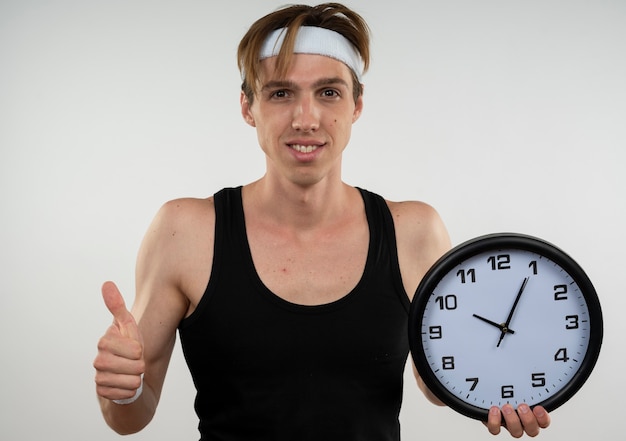 Sonriente joven deportista con diadema y pulsera sosteniendo el reloj de pared mostrando el pulgar hacia arriba aislado en blanco