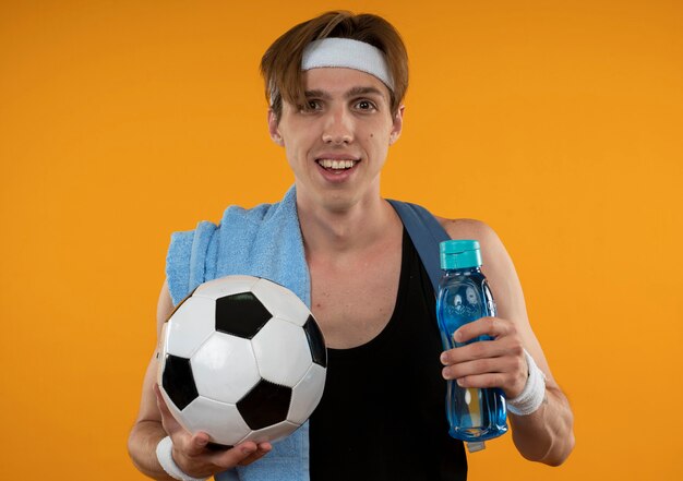 Sonriente joven deportista con mochila con diadema y muñequera con toalla en el hombro sosteniendo una botella de agua con bola aislada en la pared naranja