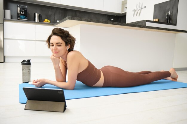 Foto gratuita sonriente joven deportista recostada sobre una estera de yoga mirando una tableta viendo un tutorial de entrenamiento físico yo