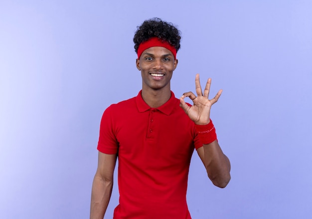Sonriente joven deportivo afroamericano con diadema y muñequera mostrando gesto okey aislado en azul