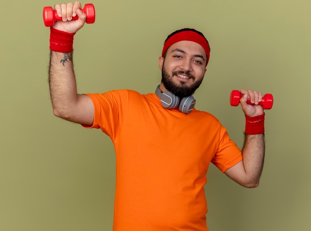 Sonriente joven deportivo con diadema y muñequera con auriculares haciendo ejercicio con pesas aislado sobre fondo verde oliva