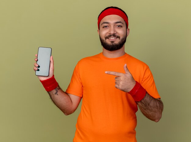 Sonriente joven deportivo con diadema y muñequera sosteniendo y apuntando al teléfono aislado en verde oliva