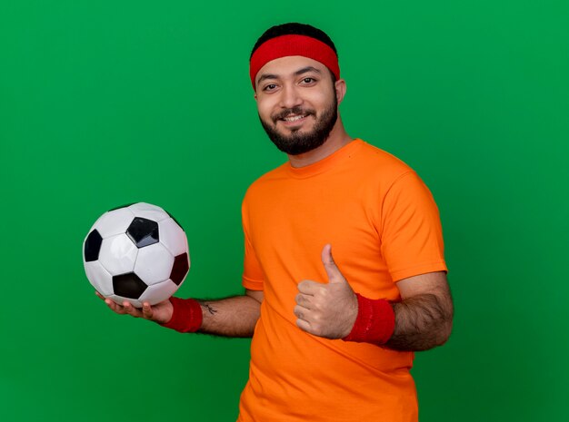 Sonriente joven deportivo con diadema y muñequera sosteniendo la bola a un lado y mostrando el pulgar hacia arriba aislado sobre fondo verde