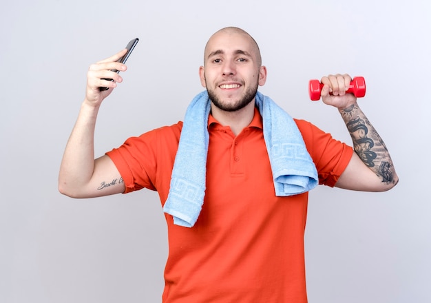 Sonriente joven deportivo levantando pesas con teléfono con toalla en el hombro aislado en la pared blanca