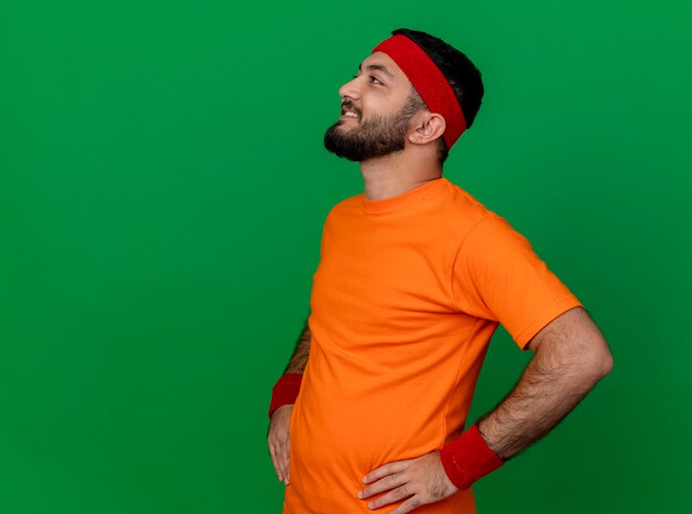 Sonriente joven deportivo mirando al lado con diadema y muñequera poniendo las manos en la cadera aislado sobre fondo verde con espacio de copia