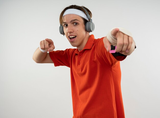 Sonriente joven deportivo vistiendo diadema con muñequera y auriculares con brazalete de teléfono que le muestra gesto