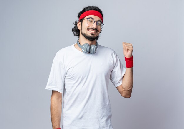 Sonriente joven deportivo vistiendo diadema con muñequera y auriculares en el cuello mostrando gesto de sí