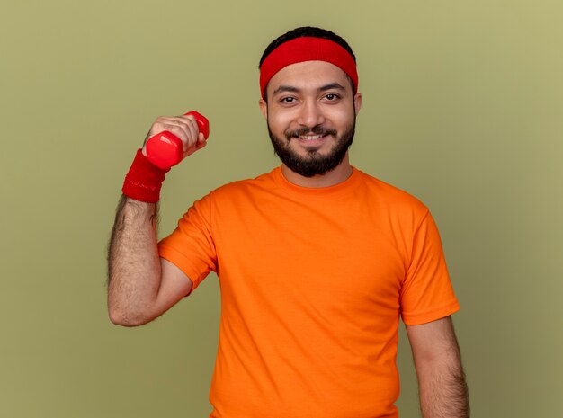 Sonriente joven deportivo vistiendo diadema y muñequera ejercicio con mancuernas aislado sobre fondo verde oliva