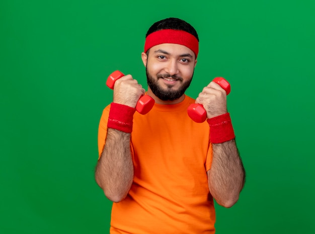 Sonriente joven deportivo vistiendo diadema y muñequera ejercicio con pesas aislado sobre fondo verde