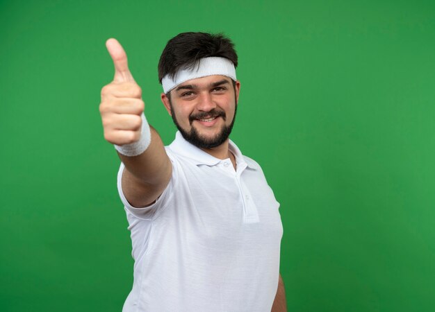 Sonriente joven deportivo vistiendo diadema y muñequera mostrando el pulgar hacia arriba aislado en la pared verde