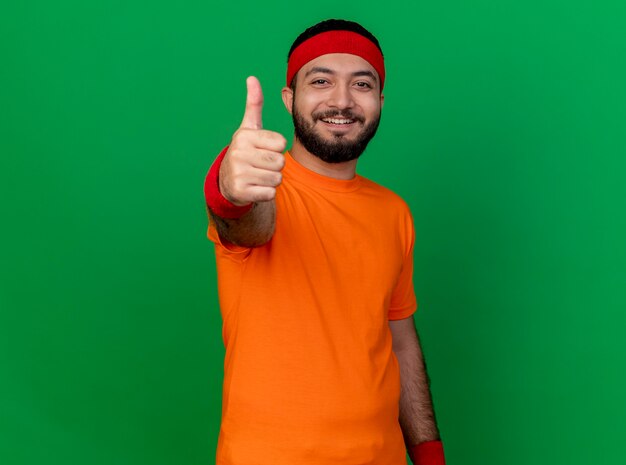 Sonriente joven deportivo vistiendo diadema y muñequera mostrando el pulgar hacia arriba aislado sobre fondo verde