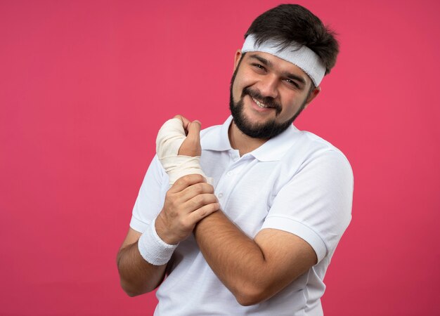 Sonriente joven deportivo vistiendo diadema y muñequera con la muñeca envuelta con una venda agarró la muñeca dolorida