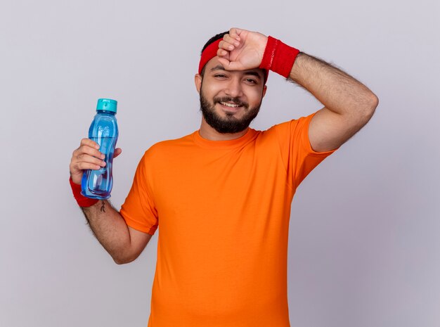 Sonriente joven deportivo vistiendo diadema y muñequera sosteniendo una botella de agua y secándose la frente con la mano aislada sobre fondo blanco.