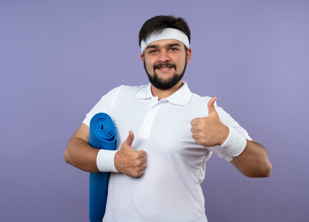 Sonriente joven deportivo vistiendo diadema y muñequera sosteniendo estera de yoga mostrando los pulgares para arriba