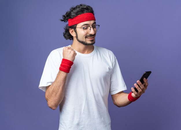 Sonriente joven deportivo vistiendo diadema con muñequera sosteniendo y mirando el teléfono mostrando sí gesto