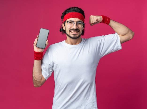 Sonriente joven deportivo vistiendo diadema con muñequera sosteniendo el teléfono mostrando un gesto fuerte
