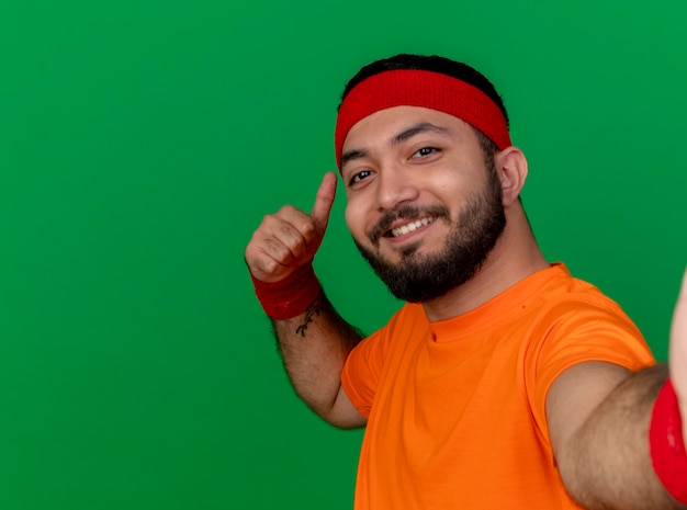 Sonriente joven deportivo vistiendo diadema y pulsera sosteniendo la cámara