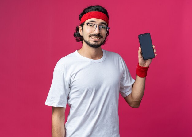 Sonriente joven deportivo vistiendo diadema con pulsera sosteniendo teléfono