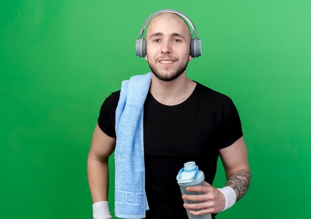 Sonriente joven deportivo vistiendo muñequera y auriculares sosteniendo una botella de agua con una toalla en el hombro aislado en la pared verde