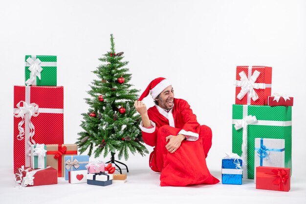 Sonriente joven divertido vestido como Santa Claus con regalos y árbol de Navidad decorado sentado en el suelo sobre fondo blanco.