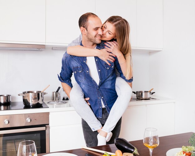 Sonriente joven divirtiéndose y dando a cuestas a su alegre esposa en la cocina