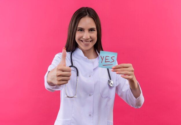 sonriente, joven, doctor, niña, llevando, estetoscopio, médico, bata, tenencia, papel, yesmark, ella, pulgar, arriba, en, aislado, pared rosa