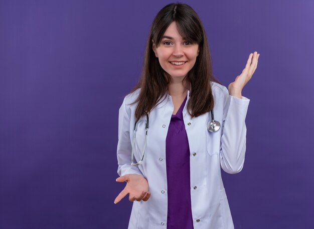 Sonriente joven doctora en bata médica con estetoscopio apunta hacia abajo y levanta la mano sobre fondo violeta aislado con espacio de copia