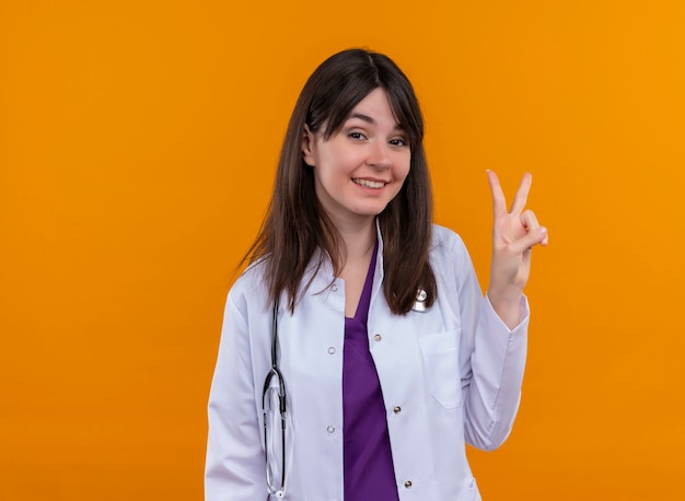 Sonriente joven doctora en bata médica con estetoscopio gestos signo de victoria sobre fondo naranja aislado con espacio de copia