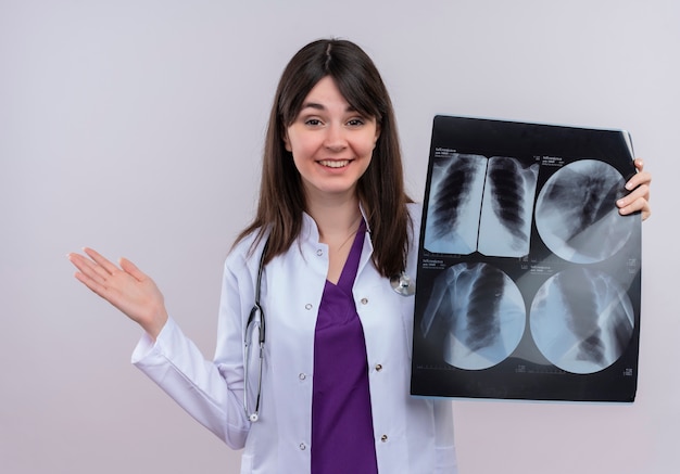 Sonriente joven doctora en bata médica con estetoscopio mantiene la mano vacía hacia arriba y sostiene la toma de rayos x sobre fondo blanco aislado con espacio de copia