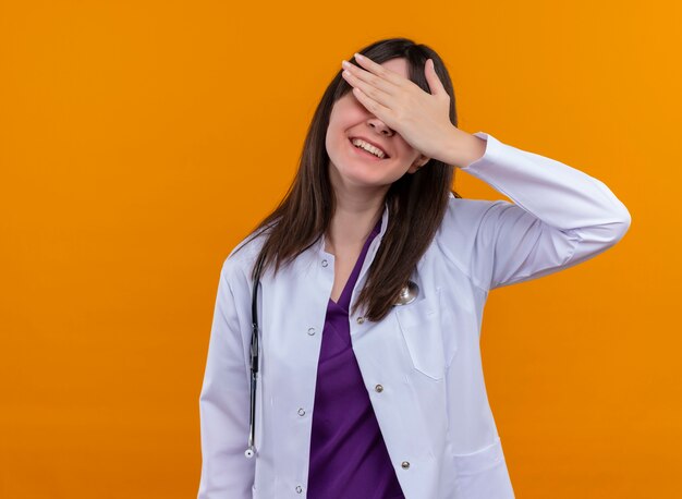 Sonriente joven doctora en bata médica con estetoscopio pone la mano en los ojos sobre fondo naranja aislado con espacio de copia