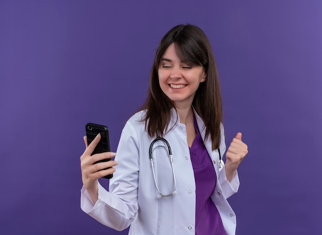 Sonriente joven doctora en bata médica con estetoscopio sostiene el teléfono y mantiene el puño en alto sobre fondo violeta aislado con espacio de copia