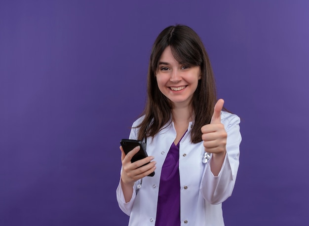 Sonriente joven doctora en bata médica con estetoscopio sostiene teléfono y pulgar hacia arriba sobre fondo violeta aislado con espacio de copia