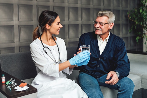 Sonriente joven doctora entrega un vaso de agua a un hombre mayor, ayudando y cuidando a una persona mayor