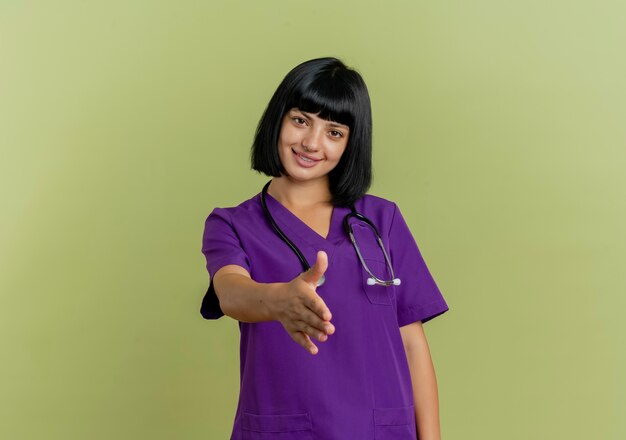 Sonriente joven doctora morena en uniforme con estetoscopio extiende la mano