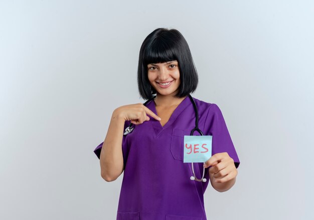 Sonriente joven doctora morena en uniforme con estetoscopio sostiene sí