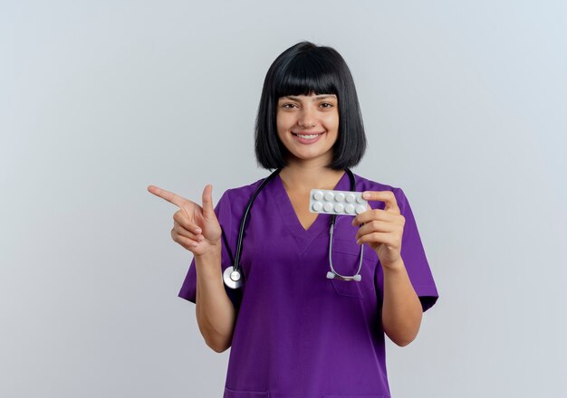 Sonriente joven doctora morena en uniforme con estetoscopio tiene medicina y puntos al lado