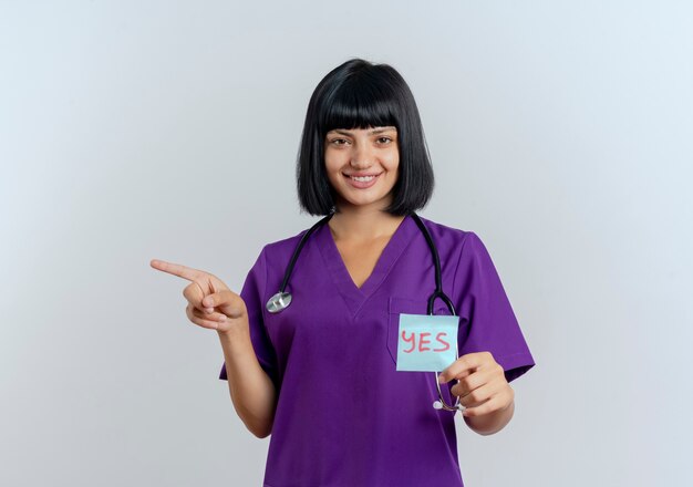 Sonriente joven doctora morena en uniforme con puntos de estetoscopio al lado