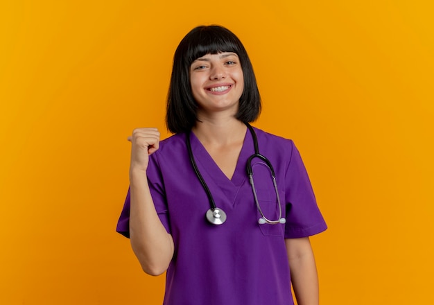 Sonriente joven doctora morena en uniforme con puntos de estetoscopio detrás aislado sobre fondo naranja con espacio de copia