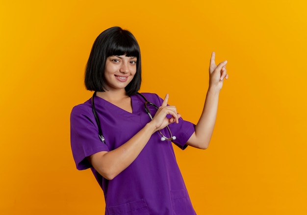 Sonriente joven doctora morena en uniforme con puntos de estetoscopio detrás con las dos manos