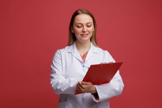 Sonriente joven doctora rubia vistiendo bata médica y estetoscopio alrededor del cuello escribiendo con lápiz en el portapapeles