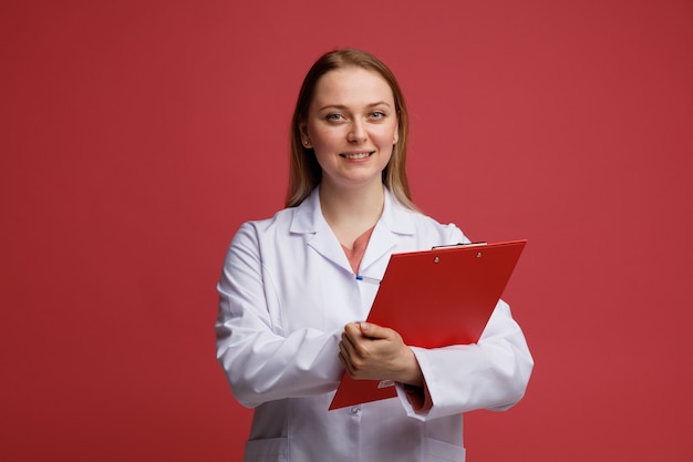 Sonriente joven doctora rubia vistiendo bata médica y un estetoscopio alrededor del cuello escribiendo en el portapapeles con lápiz