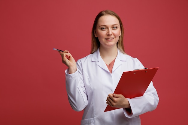 Sonriente joven doctora rubia vistiendo bata médica y un estetoscopio alrededor del cuello sosteniendo el portapapeles apuntando al lado con la pluma