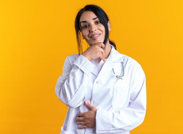 Sonriente joven doctora vistiendo bata médica con estetoscopio agarró la barbilla aislado sobre fondo amarillo